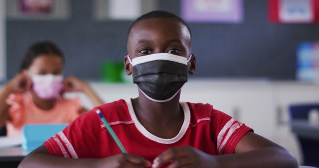 Portrait of african american schoolboy wearing face mask, sitting in classroom looking at camera. children in primary school during coronavirus covid 19 pandemic.