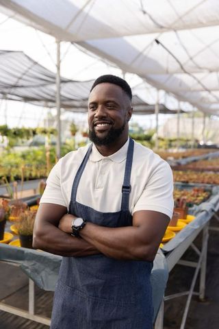 Confident african american nursery owner standing in greenhouse wearing apron and polo