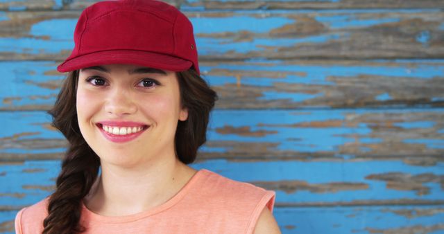 A young Caucasian woman smiles brightly, wearing a red cap against a rustic blue wooden background, with copy space. Her casual attire and cheerful expression convey a sense of relaxed, youthful style.