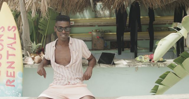 Man enjoying relaxing at tropical beach hut bar, wearing casual summer outfit and sunglasses. Perfect for themes of vacation, leisure, summer travel, tropical destinations, and beach culture marketing materials.