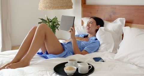 Young Woman Laughing While Using Tablet in Cozy Bedroom