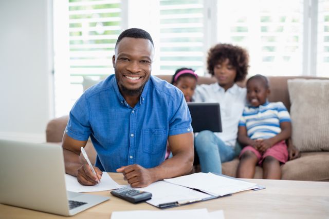 This image depicts a man smiling while calculating bills at home, with his wife and kids sitting on the sofa in the background. Ideal for use in articles or advertisements related to family finances, budgeting, home management, or work-life balance.