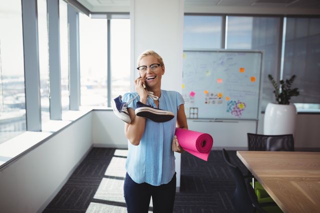 Smiling executive talking on mobile phone while holding exercise mat and shoes in office