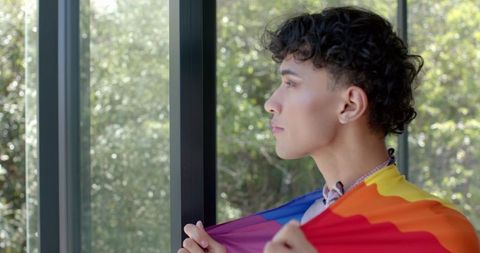 Contemplative Young Man Holding Pride Flag by Window