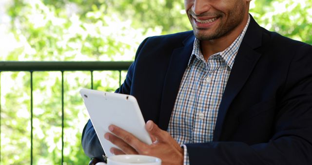 African American businessman in a suit is using a tablet outdoors, with copy space. His cheerful expression suggests he is engaged in a pleasant digital interaction or has received good news.