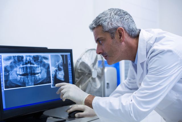 Dentist in white coat and gloves closely examining dental x-ray images on a computer monitor in a clinic. Useful for illustrating dental care, medical diagnostics, healthcare technology, and professional dental services.
