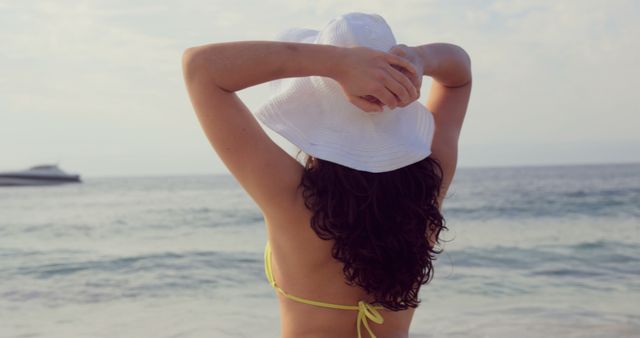 A woman in a yellow bikini, standing at the beach, adjusting her white sun hat, facing the ocean. Suitable for use in summer vacation articles, travel brochures, beachwear advertisements, and lifestyle blogs focusing on leisure and relaxation by the sea.