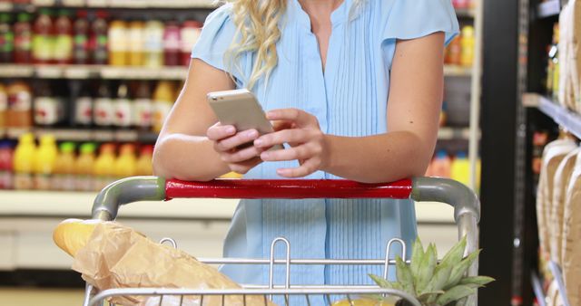 Blonde woman pushing a shopping cart in a grocery store, using a smartphone. The cart has a baguette and pineapple among other items. Ideal for themes on consumer behavior, retail experience, or modern shopping habits.