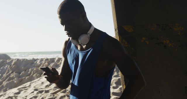African american man checking smartphone taking break during exercise outdoors by the sea. fitness, healthy and active lifestyle concept.