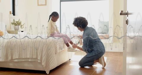 Parent kneeling tying child's sneaker on bed in cozy sunlit minimalist bedroom