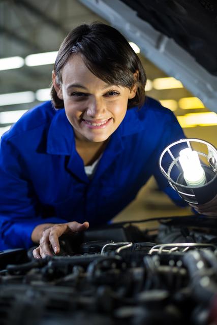 Female mechanic wearing blue uniform examining car engine with a lamp in a repair garage. She is smiling and appears focused on her work. Ideal for use in automotive repair advertisements, mechanic training materials, and promotional content for car service centers.