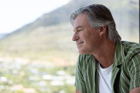 Mature man gazing out window at sunlit valley, contemplative lifestyle portrait for retirement
