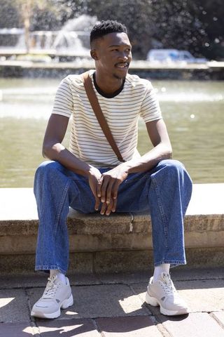 Young man sitting at plaza fountain wearing striped tee, denim jeans and white sneakers