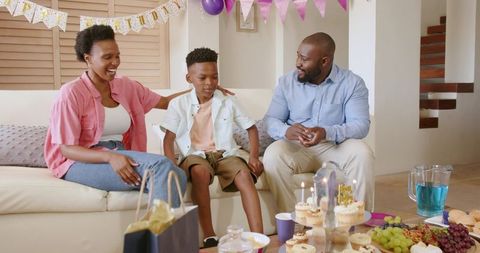 African American Family Celebrating Son's Birthday on Sofa with Cupcakes, Balloons, Candles