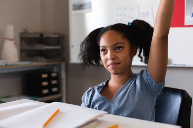 Young biracial schoolgirl sitting at a desk in a classroom, smiling and raising her hand. Ideal for educational materials, school websites, and articles about student engagement and participation. Perfect for illustrating concepts of learning, intelligence, and classroom activities.