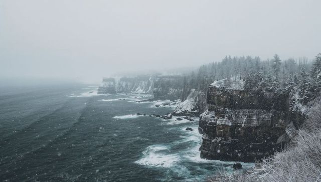 Snow-covered coastal cliffs overlooking choppy sea in winter fog with falling snow