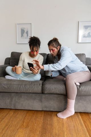 Friends relaxing on sofa sharing smartphone and coffee during cozy self-care evening