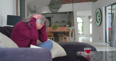 Mature woman clutching head on sofa wearing maroon sweater in modern living room