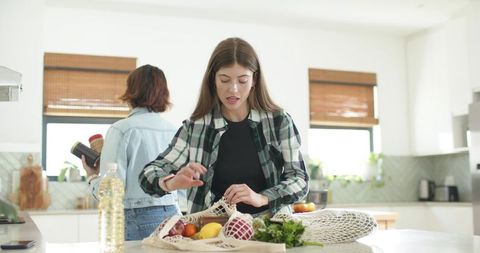 Diverse friends unpacking groceries in sunlit kitchen organizing produce into reusable mesh bags