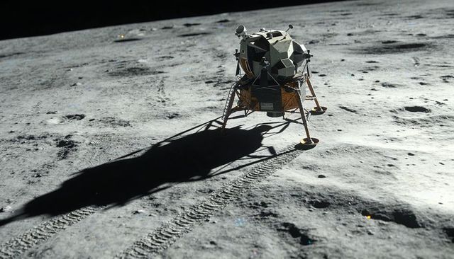 Resting lunar lander casting long shadow over moon surface with ladder and tracks
