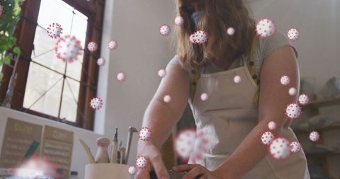Woman potter wearing mask shaping clay at studio bench with virus particle overlay