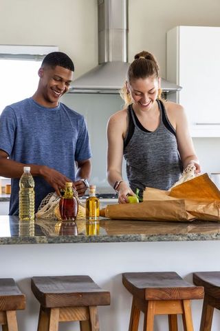 Couple unpacking groceries on sunlit modern kitchen island, smiling and preparing produce