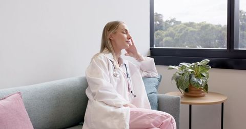 Female physician taking quiet break on clinic sofa gazing out window in sunlit lounge