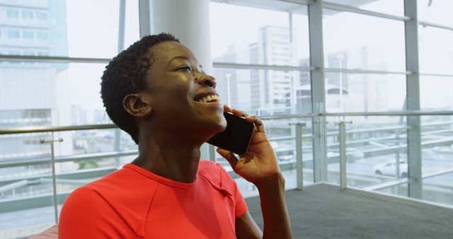 Woman with a short haircut in a red top smiling while talking on the phone in a modern office setting with large windows offering a view of the city. This stock photo can be used for advertisements, office and communication themes, or illustrating modern business environments and work-life balance.