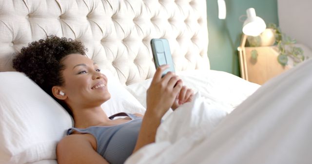 Woman lying in bed smiling and holding a smartphone, wearing earbuds. Ideal for depicting relaxation, modern technology use, morning routines, and comfortable home settings.