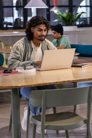 Indian man in twenties studying and working on laptop at coworking table with headphones