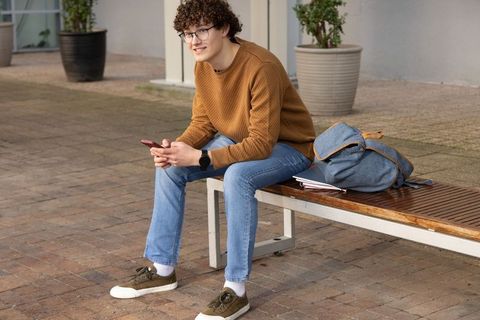 Young student sitting on bench using smartphone, backpack and notebooks in urban courtyard