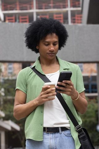 African american commuter standing in city plaza holding coffee while checking smartphone