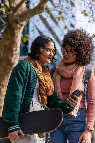 Two diverse young women checking phone and carrying skateboard on urban campus, wearing scarves
