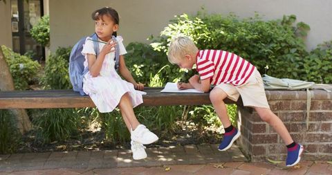 Elementary classmates sitting on bench in sunlit courtyard, boy writing, backpack scene