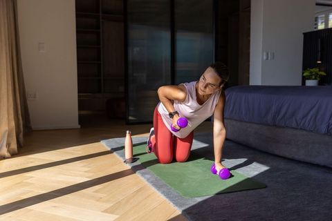 Woman kneeling on yoga mat lifting dumbbells in sunlit bedroom for home strength training