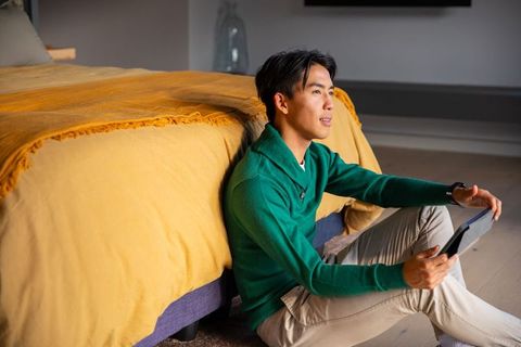 Asian man sitting on floor holding tablet wearing green sweater beside mustard bedspread