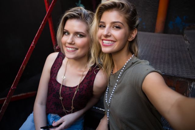 Two young women are sitting on a staircase outside a bar, smiling and enjoying their time together. This image can be used for promoting friendship, social gatherings, nightlife, and casual outings. It is ideal for use in lifestyle blogs, social media posts, and advertisements targeting young adults.