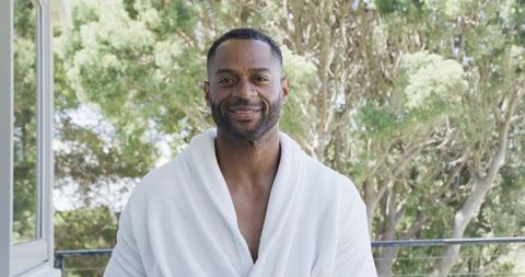 Mature man wearing white bathrobe smiling on balcony overlooking lush greenery