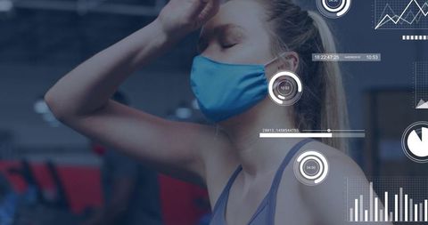 Young woman pausing at gym wearing blue face mask holding head with fitness hud metrics