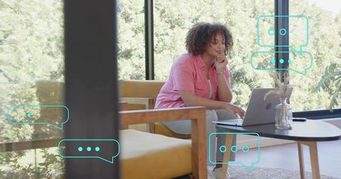 Woman typing on laptop in sunlit home workspace with chat bubbles for remote communication