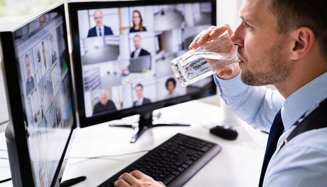 Drinking businessman attending virtual conference on dual monitors while working at modern desk