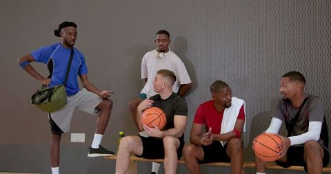 Diverse male teammates resting on bench after basketball game holding balls and gear