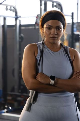 Determined asian woman athlete standing with crossed arms holding jump rope and smartwatch in gym