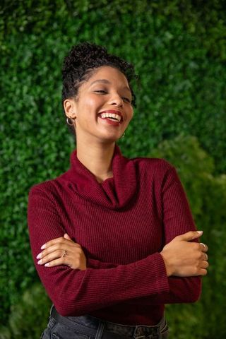 African american woman smiling and posing in burgundy sweater against green boxwood hedge