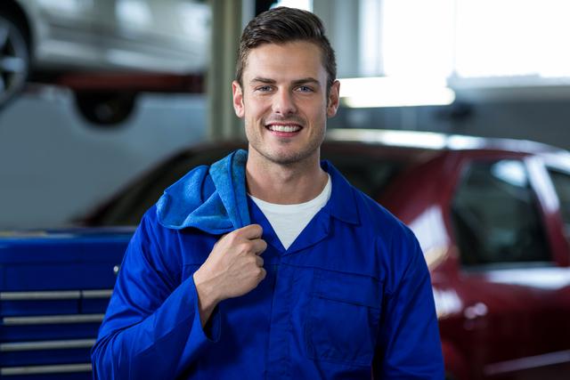 Mechanic in blue overalls smiling confidently in an auto repair garage. Ideal for use in advertisements, websites, and brochures related to automotive services, car maintenance, and professional repair services. Can also be used in articles or blogs discussing careers in the automotive industry or showcasing skilled trades.