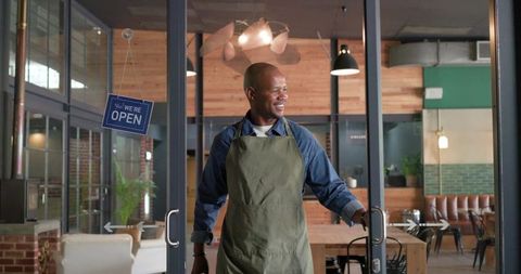 African man opening cafe doors wearing olive apron welcoming customers in rustic coffee shop
