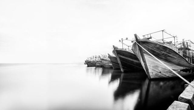 Old fishing boats are moored in a peaceful harbor. The use of black and white creates a timeless quality. Reflections of the boats in the calm water add serene beauty. This image can be used in travel magazines, for maritime-themed designs, or as wall art focusing on simplicity and tranquility.