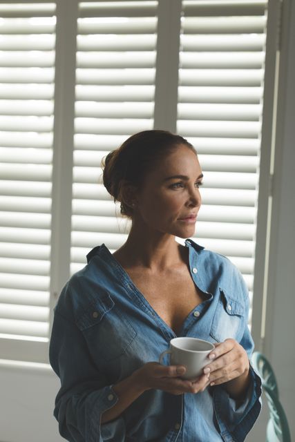 Thoughtful woman standing in living room while having coffee in a comfortable home