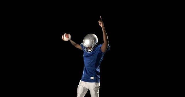 American football player celebrating with football while pointing up, wearing blue jersey and helmet. Perfect for use in sports promotions, team spirit campaigns, football events, athletic achievements, motivational posters, and fitness advertisements.