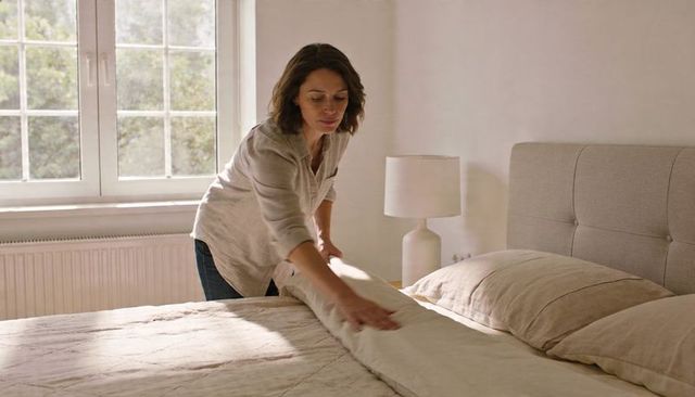 Woman making sunlit neutral linen bed in minimalist bedroom, smoothing duvet soft morning